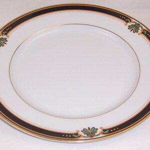 May include: A round white porcelain dinner plate featuring a wide black rim border accented with thin gold bands. The border is decorated with repeating teal and gold floral motifs and small dots, creating a symmetrical pattern around the edge.