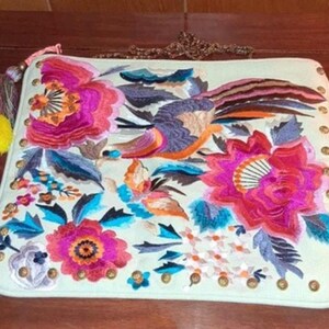 May include: A rectangular off-white clutch bag featuring vibrant floral and bird embroidery in shades of pink, orange, blue, and purple. The bag includes a metallic chain strap, gold-tone studs along the edges, and a multi-colored tassel attachment.