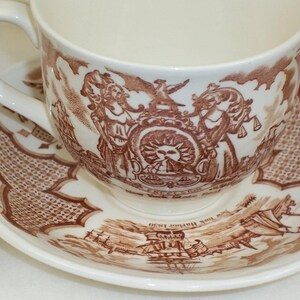 May include: A cream-colored ceramic teacup and saucer set featuring brown transferware illustrations. The cup displays the New York State Seal with the text Excelsior, while the saucer depicts a sailing ship labeled New York Harbor 1830.