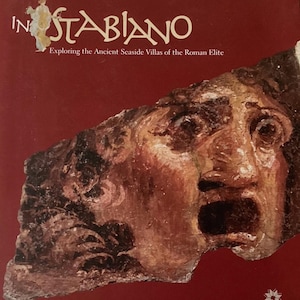 May include: A book cover with a deep red background featuring a fragment of an ancient Roman fresco depicting a face with an open mouth. The title reads IN STABIANO Exploring the Ancient Seaside Villas of the Roman Elite in white text.