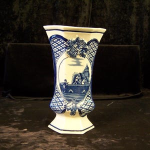 May include: A white ceramic vase featuring a blue transferware pattern of a rural landscape with a building and a figure. The vase has a flared rim, a hexagonal base, and raised relief details of grapes and leaves at the top and bottom.