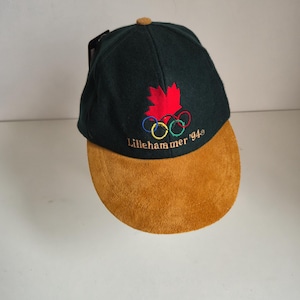 Op de afbeelding: Een donkergroene honkbalpet met een beige suède klep en een kleine beige knoop aan de bovenkant. De voorkant is voorzien van een geborduurd rood esdoornblad boven de vijf olympische ringen en de goud geborduurde tekst Lillehammer '94.