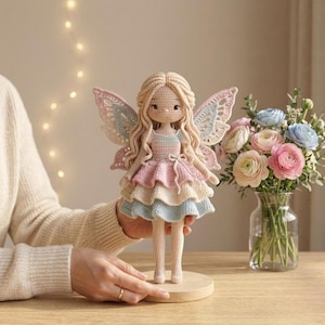May include: A handcrafted crochet fairy doll with long blonde hair and detailed wings. The doll is dressed in a layered pastel dress of pink, blue, and cream. The doll stands on a small wooden base. A bouquet of flowers is in the background.