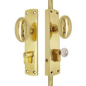 May include: A polished brass door hardware set featuring two oval-shaped knobs mounted on rectangular backplates. The assembly includes a cylinder lock mechanism on each plate, with a small silver key inserted into the right-side lock cylinder.