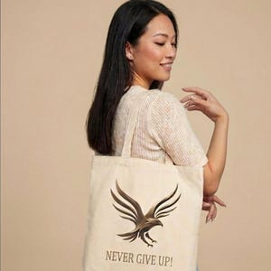 May include: A beige canvas tote bag featuring a dark brown graphic of a soaring eagle with its wings spread wide. Below the eagle illustration, the text "NEVER GIVE UP!" is printed in a simple, dark brown sans-serif font on the front of the bag.