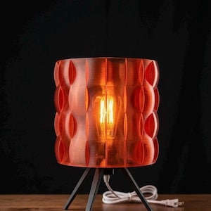 May include: A table lamp featuring a cylindrical, translucent orange shade with a repeating vertical oval pattern. The lamp is supported by three thin, black metal legs and contains a glowing Edison-style light bulb with a visible white power cord.