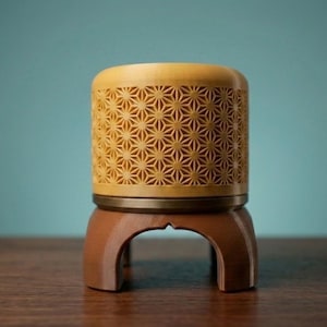 Puede incluir: Un recipiente decorativo de madera clara con un patrón geométrico de estrellas tallado en los lados. El recipiente es de color marrón claro y se apoya sobre un soporte de tres patas de color marrón oscuro. El fondo es turquesa. Aproximadamente 10 cm de altura.