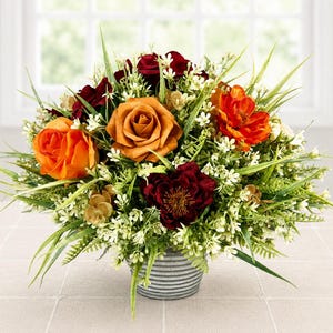 May include: A floral arrangement featuring orange, rust, and deep burgundy roses and flowers mixed with green grass and small white filler blossoms. The bouquet is contained in a small, round, grey metal planter with horizontal ribbed detailing.