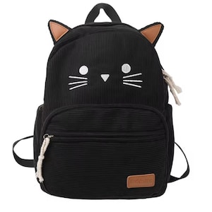 May include: Black corduroy backpack with cat face details. Features embroidered white whiskers, ear accents, and a front pocket. Includes a top handle and adjustable straps. The backpack has a small brown rectangular tag with text.