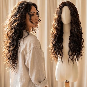 May include: A woman with long, wavy, dark brown hair stands next to a wig on a mannequin. The wig has a similar style and color to the woman's natural hair. The hair is styled with loose waves, and the mannequin is on a wooden stand.