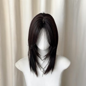 May include: A layered wig with dark brown and black highlights, styled with face-framing layers. The wig is displayed on a white mannequin head, with a silver necklace visible. The hair has a natural-looking texture and a subtle shine.