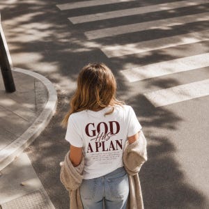 Op de afbeelding: Een wit T-shirt met korte mouwen en de donkerrode tekst GOD HAS A PLAN op de achterkant, met daaronder kleinere bijbeltekst. Het shirt wordt gedragen met een blauwe spijkerbroek en een beige vest over de schouders.