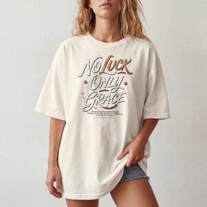 Op de afbeelding: Een gebroken wit t-shirt met korte mouwen en de tekst "NO LUCK ONLY GRACE" in een gestileerd bruin en wit lettertype. Onder de hoofdtekst staat kleinere tekst: "FOR BY GRACE YOU HAVE BEEN SAVED THROUGH FAITH, AND THAT NOT OF YOURSELVES: IT IS THE GIFT OF GOD. EPHESIANS 2:8".