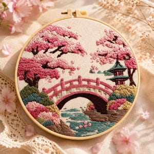 May include: Hand-stitched embroidery art in a wooden hoop. The design features a Japanese landscape with a pink bridge, cherry blossom trees, and a pagoda. The color palette includes shades of pink, green, brown, and cream.