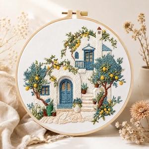 Op de afbeelding: Een ronde borduurring met een wit stoffen paneel met een gedetailleerde geborduurde scène van een wit huis in mediterrane stijl met blauwe deuren en ramen, omringd door groene bomen met gele citroenen en een stenen trap.