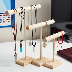 May include: Three light-colored wooden T-bar jewelry display stands of varying heights. The stands hold several necklaces and a red cord bracelet. The jewelry includes a silver wing pendant, a blue and brown resin pendant, and a white horn-shaped charm.