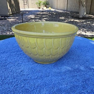 May include: A large, yellow ceramic bowl with a textured, scale-like pattern. The bowl has a wide rim and sits on a blue surface. The bowl is likely for decorative or functional use.
