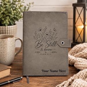 May include: A grey notebook with a snap closure featuring a laser-engraved floral design, a butterfly, and the text "Be Still & know PSALM 46:10" centred on the front cover. The bottom of the cover includes the placeholder text "Your Name Here".