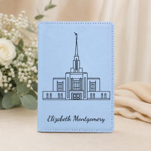 May include: A light blue passport holder featuring a black line art illustration of a temple with a tall spire and the name Elizabeth Montgomery printed in a cursive script font below the building design on the front cover.