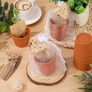 May include: Small terracotta pots with seed packets, some in sheer bags, and heart-shaped tags that read "Thank you" and "Let Love Grow." A cup of coffee and decorative greenery are also present.