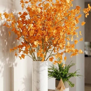 May include: A tall cylindrical vase with a white marble finish containing a large arrangement of vibrant orange flowers with delicate petals and thin green stems, positioned next to a small green potted plant and a wooden triangular ornament.