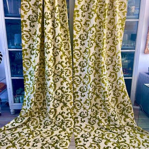 Puede incluir: Dos cortinas color crema con un patrón repetido de volutas florales verdes estilizadas. Las cortinas cuelgan frente a un armario blanco con puertas de vidrio. La tela cae hasta el suelo.
