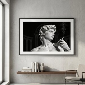 May include: Black and white framed print featuring a marble statue of David holding a cigarette, with smoke rising. The background is black with architectural line drawings. The print is displayed above a wooden shelf with books and a vase.