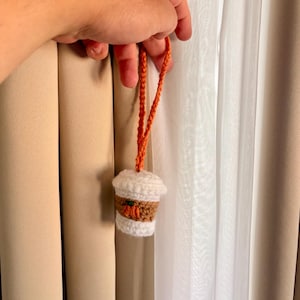 May include: A small, crocheted coffee cup ornament featuring a white lid, a tan middle section with a tiny orange pumpkin appliqué, and a white base. The cup is attached to a thin, orange crocheted loop for hanging, crafted with yarn.
