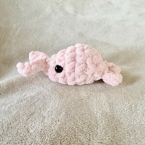 May include: A handmade, light pink crochet whale plushie with a black eye. The soft, textured toy is lying on a light gray, textured surface. The whale has a rounded body and small fins.