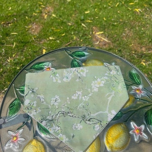 Op de afbeelding: Een bandana met een groen en wit bloemenpatroon, geplaatst op een glazen serveerschaal met citroen- en bladmotieven. De schaal staat op gras.