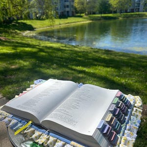 May include: An open book with colorful tabs, resting on a round glass table. The book is surrounded by a decorative fabric with a lemon and floral pattern. The background features a lake and green landscape.