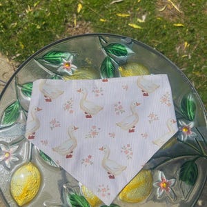 Op de afbeelding: Een witte bandana met een patroon van eenden en kleine roze bloemen. De bandana is in een driehoek gevouwen en ligt op een glazen tafel met citroen- en bloemmotieven.