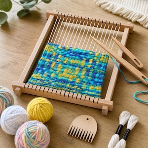 May include: A rectangular wooden weaving loom with a partially completed tapestry in shades of blue, teal, and yellow. Includes two wooden weaving needles, a wooden weaving comb, three balls of yarn, and two white thread skeins for crafting.