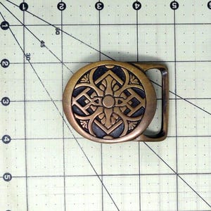 May include: An oval-shaped brass belt buckle featuring a dark recessed center with an intricate raised geometric pattern. The buckle is positioned on a cutting mat with a grid, showing it measures approximately 2.5 inches in width.