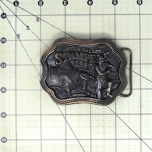 May include: A bronze-toned metal belt buckle featuring a relief design of a bull and a rider. The text reads CORN PALACE STAMPEDE 1987 MITCHELL S.D. The buckle is positioned on a cutting mat with a grid showing a width of approximately 3.5 inches.