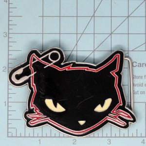 May include: A black metal belt buckle shaped like a cat head with cream-colored eyes and nose, outlined in red. A silver safety pin design is attached to the left ear. The buckle is positioned on a grid mat showing a width of 3.5 inches.