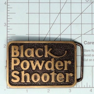 May include: A rectangular brass belt buckle with a textured dark background and raised gold-toned lettering that reads Black Powder Shooter. The buckle is positioned against a grid mat showing a width of approximately 3.5 inches and a height of 2.25 inches.