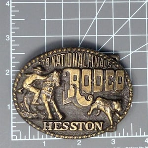 May include: An oval brass-toned belt buckle featuring a relief of a rider on a horse and a calf. The text reads '78 NATIONAL FINALS RODEO' and 'HESSTON'. A ruler visible in the background indicates the buckle is approximately 3.5 inches wide.
