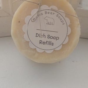 May include: A round, off-white solid dish soap bar tied with thin brown twine. A scalloped white paper label is attached to the front, featuring a bear icon and the text "Mama Bear Soaps" above the words "Dish Soap Refills" in dark ink.
