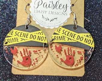 May include: A pair of black and yellow crime scene do not cross tape earrings with a red bloody handprint on a clear background.