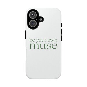 Può includere: Una custodia per smartphone bianca con un modulo fotocamera nero dotato di due lenti circolari e un piccolo flash. Il testo 'be your own muse' è stampato al centro in un carattere serif verde scuro, con 'muse' in dimensioni maggiori.