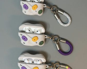 May include: Three white textured earbud cases shaped like climbing walls with purple, yellow, and green holds. Each case contains white earbuds and features a metal carabiner attached to a short, colorful rope loop in grey, purple, or multicolored.