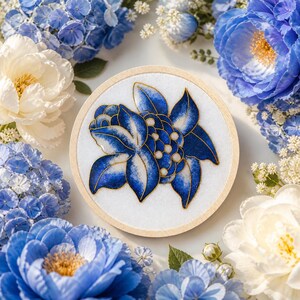Può includere: Un telaio da ricamo rotondo in legno con un motivo floreale blu e bianco e dettagli dorati. Il design è circondato da fiori bianchi e blu, creando un'esposizione decorativa e artistica. Un oggetto artigianale.