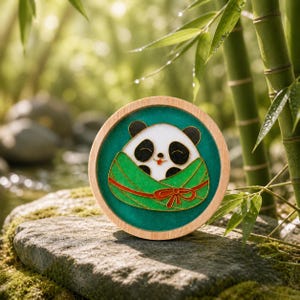 Könnte beinhalten: Ein runder Holzuntersetzer mit einem Panda-Bär-Design. Der Panda ist in eine grüne Decke mit einer roten Schleife gewickelt. Der Untersetzer hat einen türkisfarbenen Hintergrund und befindet sich vor einem natürlichen Hintergrund im Freien.