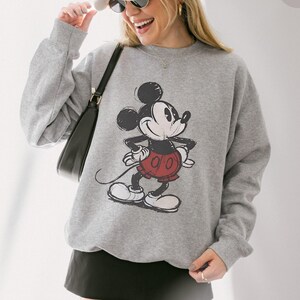 May include: Athletic heather gray sweatshirt featuring a distressed graphic of Mickey Mouse in black, red, and white. The sweatshirt has a crew neck and long sleeves. The text "ATHLETIC HEATHER" is in the upper right corner.