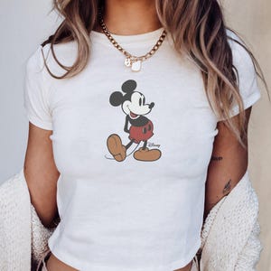 K&ouml;nnte beinhalten: Wei&szlig;es Crop-Top mit Mickey Mouse-Grafik. Die Zeichentrickfigur tr&auml;gt rote Shorts, braune Schuhe und schwarze Ohren. Das Wort "Disney" ist auf dem Shirt aufgedruckt. Das Wort "White" ist unten links aufgedruckt.