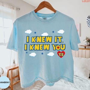 May include: A light blue short-sleeve t-shirt featuring the yellow text I KNEW IT, I KNEW YOU with a red heart containing the letters TS, surrounded by white clouds and small blue stars. The shirt is displayed on a wooden hanger.