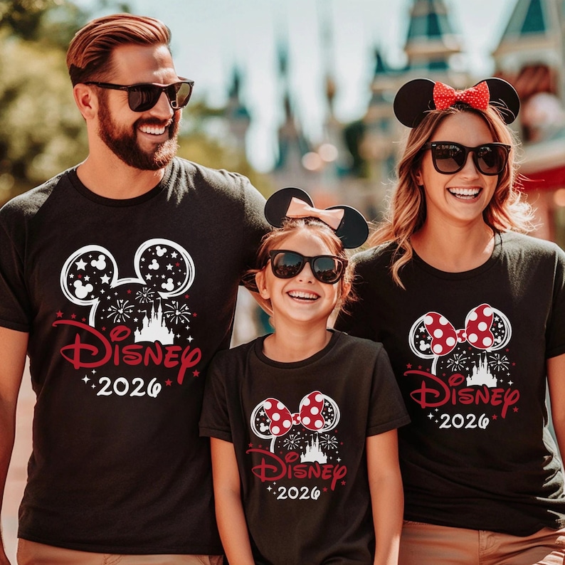 Puede incluir: Tres camisetas negras con un estampado gr&aacute;fico en blanco y rojo de una silueta de Mickey Mouse, un castillo, fuegos artificiales, estrellas, el texto Disney y el a&ntilde;o 2026. Las camisetas son usadas por tres personas que llevan gafas de sol.