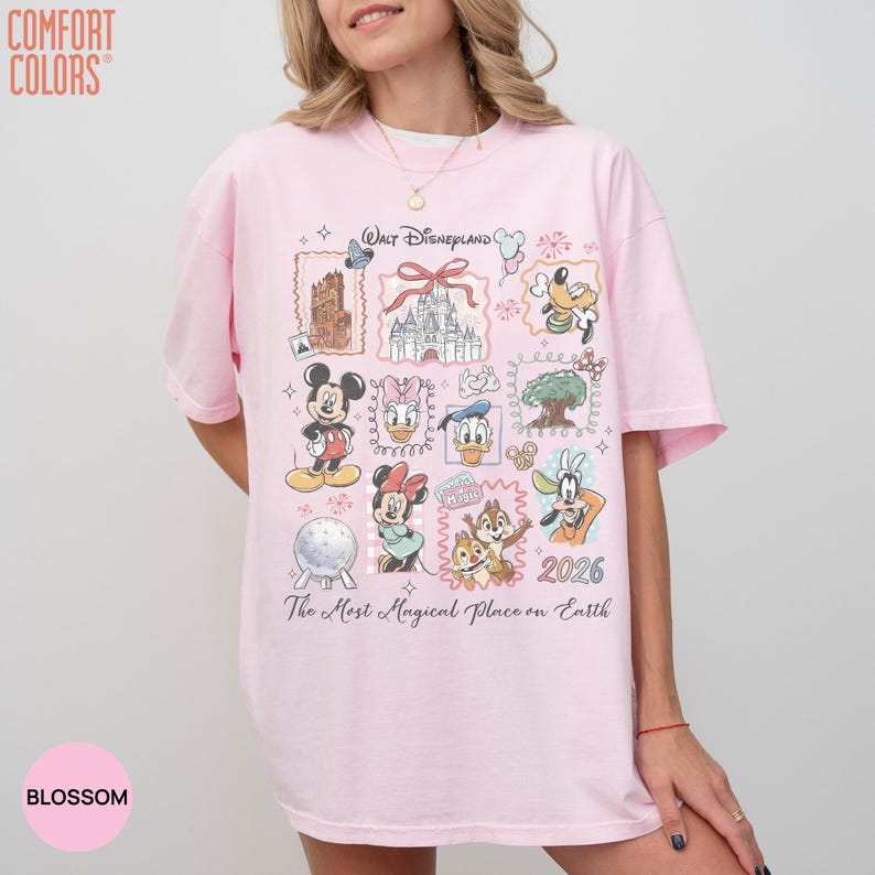 May include: A light pink t-shirt featuring a graphic print with Walt Disneyland text, a castle, and various cartoon characters including Mickey Mouse, Minnie Mouse, Donald Duck, Daisy Duck, Goofy, and Chip and Dale, with the year 2026 and The Most Magical Place on Earth text.