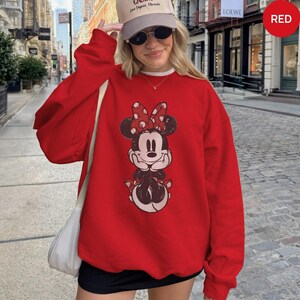 May include: A red sweatshirt featuring a Minnie Mouse graphic. The graphic shows Minnie Mouse with a red bow and a smiling face. The sweatshirt is a solid red color and has long sleeves.